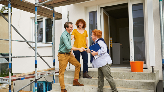 Drei Personen stehen vor einem Haus im Bau, eine Person schüttelt einer anderen die Hand, während eine dritte Person zuschaut; im Hintergrund sind ein Gerüst und ein orangefarbener Eimer zu sehen.