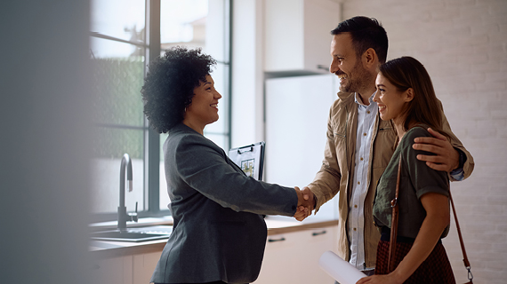 Ein Paar schüttelt einer Person in einem Anzug die Hand in einer modernen Küche. Die Person im Anzug hält eine Mappe.