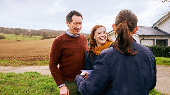 Drei Personen stehen im Freien, zwei davon einander zugewandt, während eine dritte Person ihnen gegenübersteht; im Hintergrund befinden sich ein Feld und ein Haus.