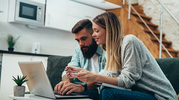 Zwei Personen sitzen auf einem Sofa und arbeiten gemeinsam an einem Laptop; im Hintergrund sind eine Küche und eine Treppe zu sehen.