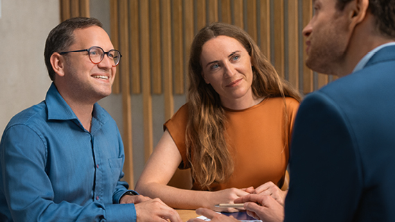 Drei Personen sitzen an einem Tisch und führen ein Gespräch; eine vierte Person ist teilweise sichtbar im Vordergrund.